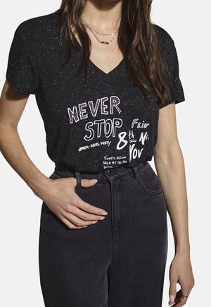 Femme portant un t-shirt noir à col en V avec des taches et l'inscription « NEVER STOP » ainsi que les détails d'un événement imprimés, associée à un jean noir taille haute.