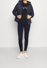 Kvinde iført en navy quiltet jakke, sort sweatshirt med hvid skrift, mørke skinny jeans og hvide sneakers, stående foran en ensfarvet baggrund.