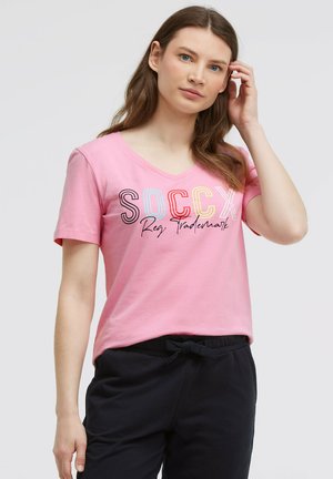 Young woman with long brown hair wearing a pink V-neck T-shirt with colorful SOCCX logo and black drawstring pants against a plain background.