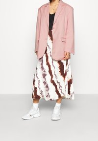 Różowa, oversize'owa marynarka nałożona na czarną bluzkę, zestawiona z midi spódnicą w stylu tie-dye w białym, brązowym i ciemnoczerwonym kolorze oraz białymi sneakersami.