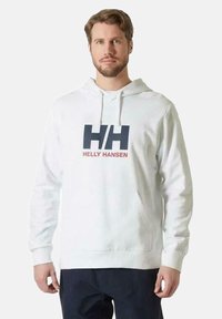 Sudadera blanca de algodón con un bolsillo canguro en la parte delantera, que presenta un gran logo "HH" y el texto "Helly Hansen" en azul marino y rojo. Capucha ajustable con cordón.
