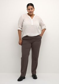 Weiße Bluse mit kleinen blauen Mustern, geknöpfter Hals, hochgerollte Ärmel, kombiniert mit maßgeschneiderten braunen Hosen und schwarzen Slippern.