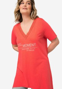 Camiseta color coral con mangas cortas, escote en V y detalles de encaje en el cuello. Presenta texto gráfico en blanco. Tejido suave.