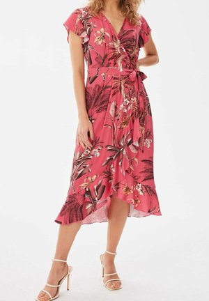 Vrouw draagt een roze bloemen wikkeljurk met korte vleermuismouwen en een gerimpelde zoom, gecombineerd met beige hoge hakken.