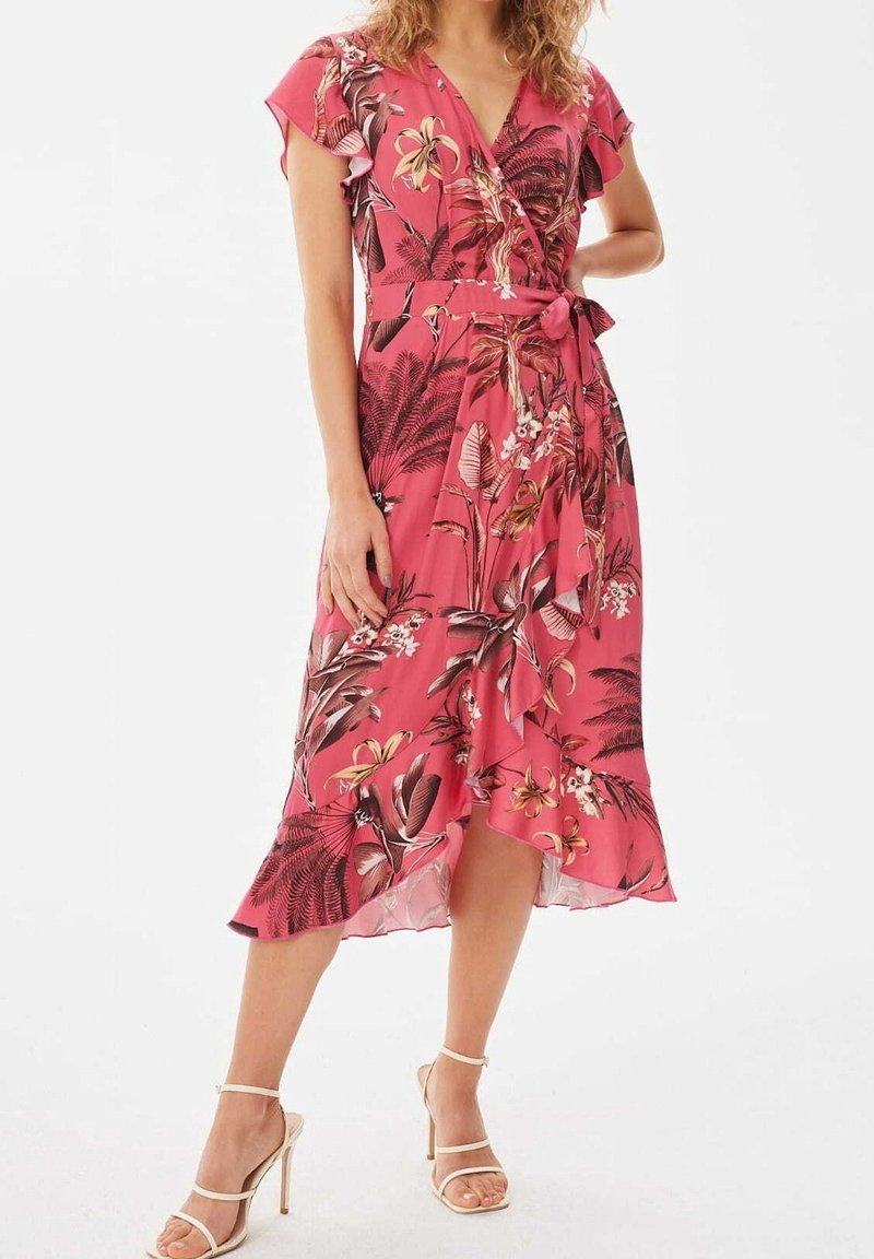 Femme portant une robe portefeuille à fleurs rose avec des manches courtes volantées et un ourlet à volants, associée à des sandales à talons hauts beiges.