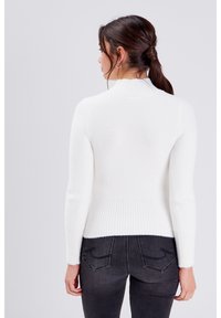 Pull en maille blanche, doux, ajusté, avec un col haut en côtes et des manches longues. Corps texturé et ourlet en côtes, porté avec un jean gris foncé.