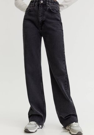 Personne portant un jean noir taille haute à jambes larges et des baskets blanches, debout devant un fond clair uni.