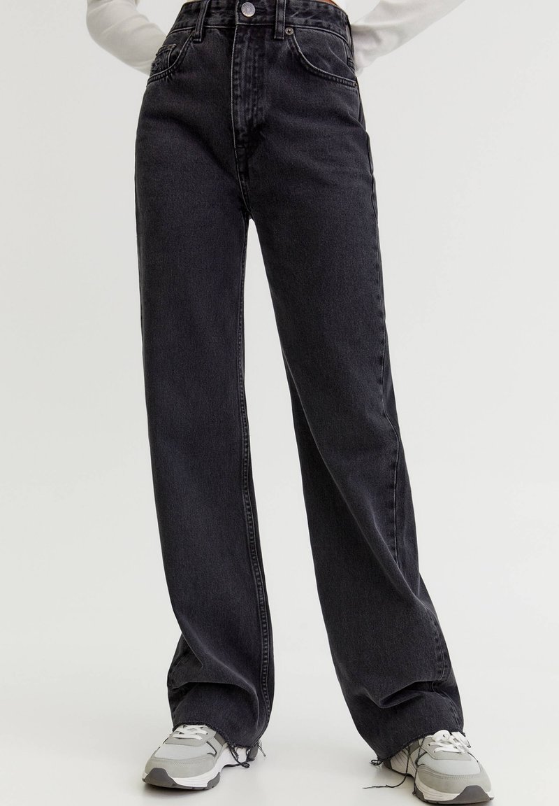 Personne portant un jean noir taille haute à jambes larges et des baskets blanches, debout devant un fond clair uni.