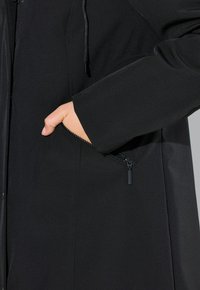 Main dans la poche zippée d'un manteau noir, avec la tirette de la fermeture éclair et la texture du tissu visibles de près.