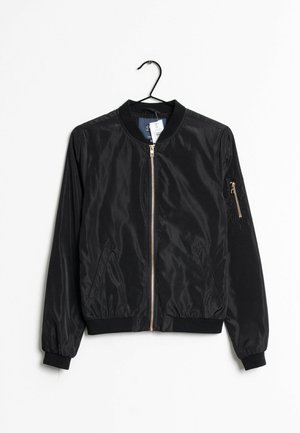 Czarna kurtka bomberka wykonana z gładkiego, lekkiego materiału. Ma zamek z przodu, ściągacze przy mankietach oraz zasuwaną kieszeń na rękawie. Wisi na haku.