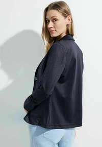 Blazer bleu marine foncé avec une texture lisse, col, fermeture à boutons, poche poitrine unique et manches longues, superposé sur un pantalon bleu clair.