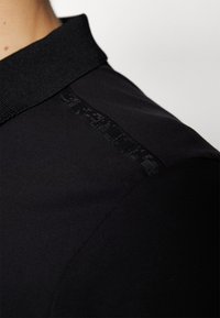 Gros plan sur l'épaule d'une chemise noire avec un col côtelé et un subtil détail de bande texturée le long de la couture de l'épaule.