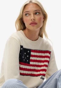 Sweter dzianinowy w odcieniu kremowym z motywem flagi amerykańskiej, z czerwonymi i granatowymi akcentami, teksturowymi pasami oraz ribbowym dekoltem.