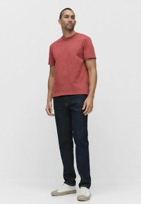 Rød bomulds t-shirt med rund hals, parret med mørkeblå denim jeans og beige sneakers. Modellen står foran en neutral baggrund.