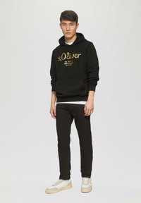 Zwarte hoodie met gouden "s.Oliver" logo, gecombineerd met zwarte broeken en lichtgekleurde sneakers, met een gladde textuur en een relaxte pasvorm.