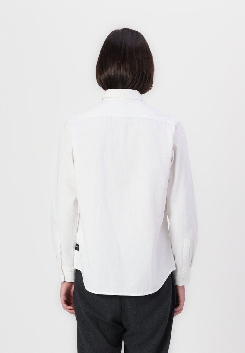 Persoon met kort donker haar, gekleed in een witte blouse met lange mouwen en donkere broeken, gefotografeerd van achteren tegen een witte achtergrond.