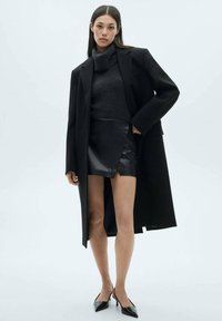 Black oversized coat over a grey turtleneck sweater, paired with a black leather mini skirt and black pointed-toe heels. Smooth textures.