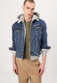 Denim jacket with shearling collar in dark blue, featuring button closures, chest pockets, and gray hoodie underlayer. Smooth texture.