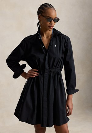 Γυναίκα με πλεγμένα μαλλιά φοράει γυαλιά ηλίου και μαύρο φόρεμα με γιακά και μακριά μανίκια, με ζώνη στη μέση, στέκεται μπροστά σε απλή φόντο.