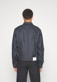 Persona con cabello rizado y corto lleva una chaqueta de mezclilla oscura y pantalones negros, de pie frente a un fondo blanco liso.