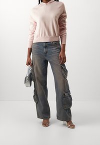 Rosa Kapuzenpullover mit Kordelzug, kombiniert mit blauen Cargo-Jeans mit Seitentaschen und transparenten Sandalen mit Absatz. Silberne Handtasche in der Hand.