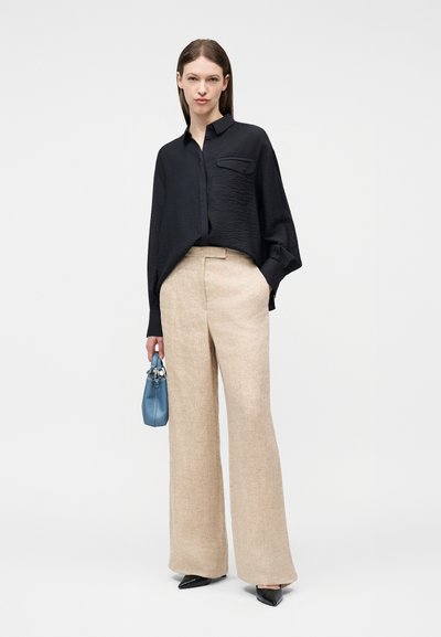 Γυναίκα που φοράει μαύρο, άνετο πουκάμισο με μακριά μανίκια, μπεζ παντελόνι φαρδύ, μαύρα μυτερά παπούτσια, κρατώντας μια μικρή μπλε τσάντα χειρός.