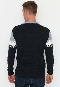 Pull en tricot bleu marine avec motif de vague, épaules grises et accents géométriques blancs et noirs sur les manches. Poignets et ourlet côtelés.