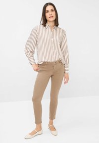 Camicia a maniche lunghe a righe beige e crema, abbinata a pantaloni aderenti color pelle. Modella che indossa scarpe basse chiare, in piedi su uno sfondo neutro.