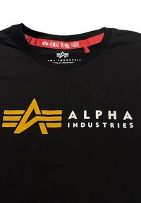 Juodas medvilninis marškinėliai su auksiniu ir baltu "ALPHA INDUSTRIES" logotipu ir stilizuotu sparno dizainu apačioje. Matomas raudonas žymeklį "REMOVE BEFORE FLIGHT".