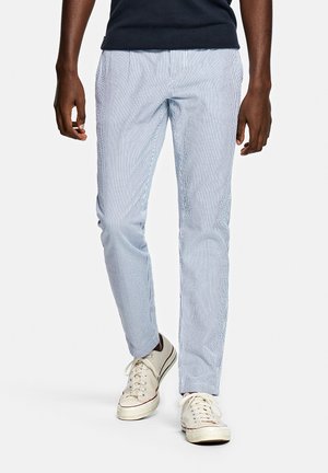 Homme portant un pantalon ajusté à rayures verticales bleu clair et blanc, avec des baskets en toile écru à lacets blancs, debout devant un fond blanc.