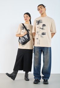 T-shirt beige oversized con stampe floreali nere; gonna di jeans nera con spacco frontale; jeans baggy blu; stivali neri; borsa a tracolla nera.