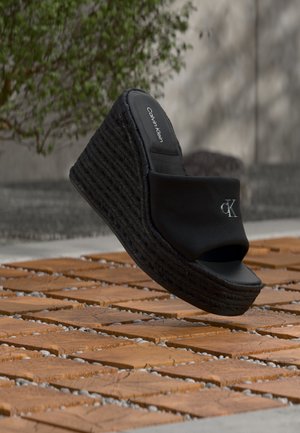 Sandalia cuña negra de Calvin Klein con suela de plataforma trenzada, puntera abierta y logotipo "cK" en la parte superior, colocada sobre un suelo de baldosas de madera al aire libre.