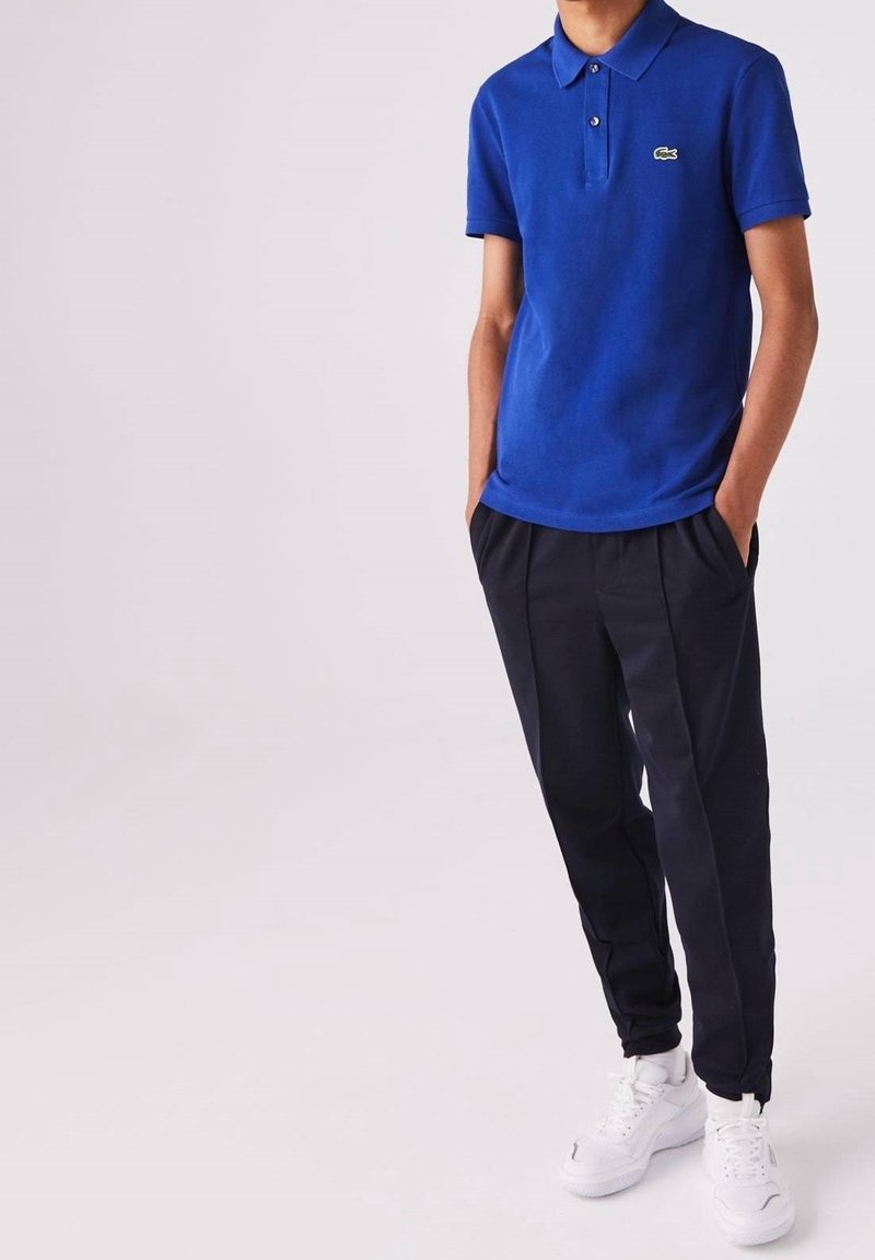 Personne portant un polo bleu avec un petit logo, un pantalon noir et des baskets blanches, debout les mains dans les poches devant un fond uni.