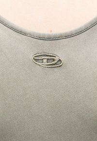 Κοντινό πλάνο στο λαιμό ενός ανοιχτό γκρι πουκαμίσου με ορατή ραφή και κεντημένο οβάλ σχέδιο ματιού λίγο πιο κάτω από το γιακά.