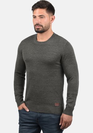 Suéter de punto gris con mangas largas, cuello redondo y puños acanalados. Presenta un pequeño parche de marca en la parte inferior izquierda.