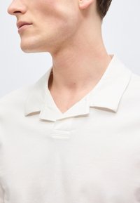 Νεαρό άτομο που φοράει λευκό πουκάμισο με γιακά, το κεφάλι γυρισμένο προς τα αριστερά, εμφανίζοντας το πλαϊνό προφίλ και το λαιμό σε απλό φόντο.