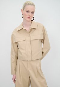 Giacca beige cropped con colletto, due tasche frontali e maniche lunghe. Tessuto liscio dal design minimalista.
