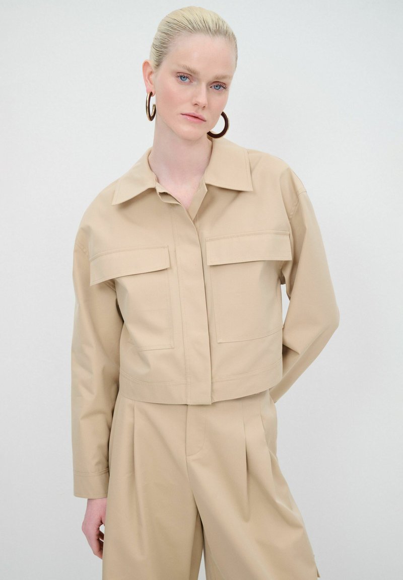 Giacca beige cropped con colletto, due tasche frontali e maniche lunghe. Tessuto liscio dal design minimalista.