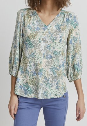 Kobieta w kwiecistej bluzce z dekoltem w serek z bufiastymi rękawami 3/4 oraz dopasowanych jasnoniebieskich spodniach na jednolitym tle.