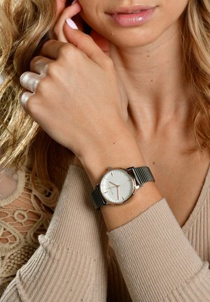 Silberne Uhr mit strukturiertem weißen Zifferblatt, schwarzem Metallnetzarmband und rundem Gehäuse. Trägt man am Handgelenk, teilweise von einer Hand verdeckt.