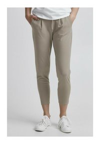 Lys beige, tapered bukser med en glat tekstur, der har forlommer og elastisk talje, parret med hvide sportsko.