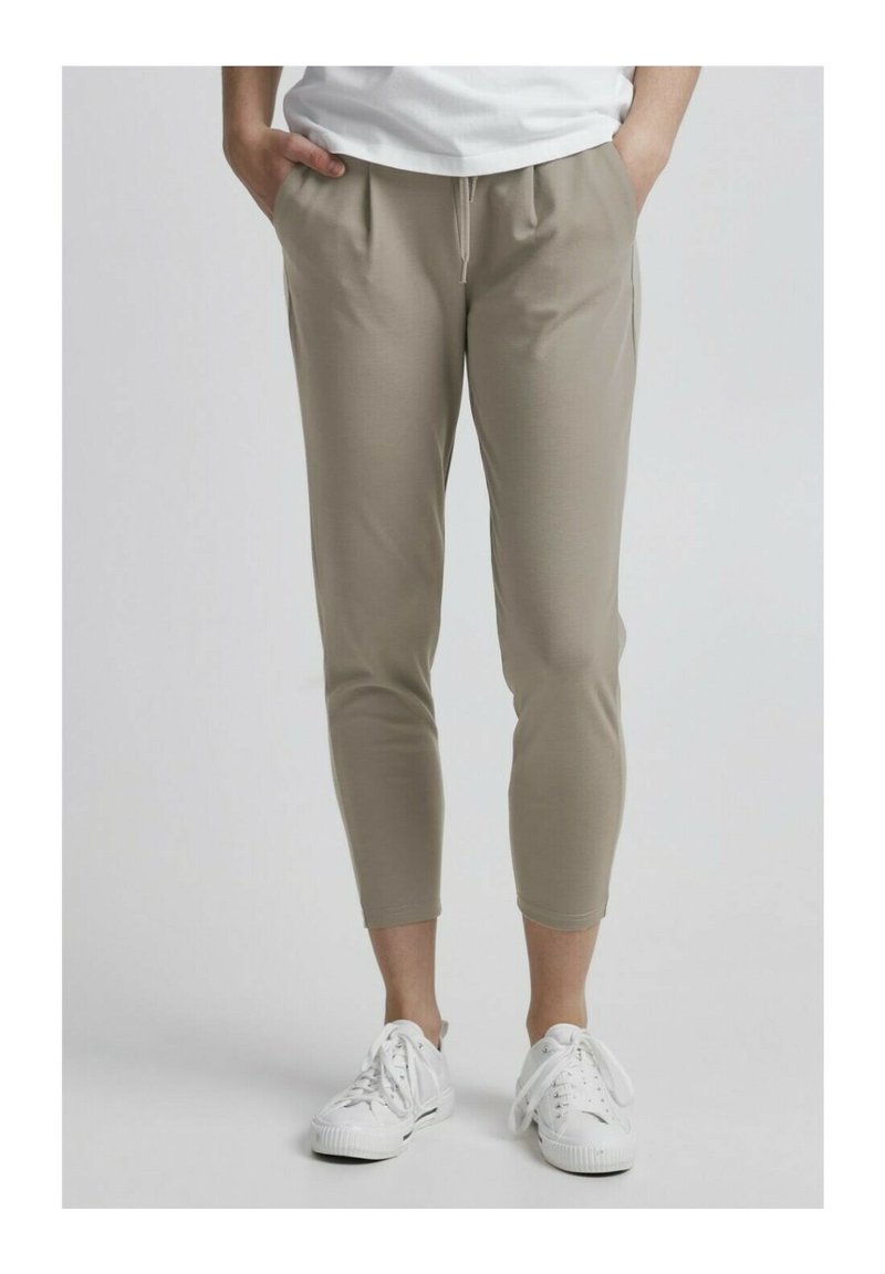 Lys beige, tapered bukser med en glat tekstur, der har forlommer og elastisk talje, parret med hvide sportsko.