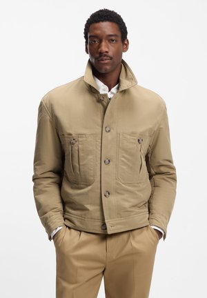 Khaki padded jacket with high collar, two chest pockets, and button closure; paired with beige trousers. Smooth texture and tailored fit.