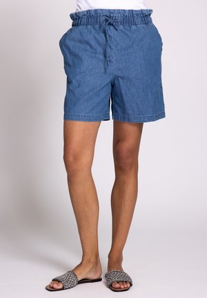 Blaue Shorts mit elastischem Bund und Kordelzug, getragen von einer stehenden Person, kombiniert mit schwarz-weißen gewebten Sandalen mit offener Zehenpartie und flacher Sohle.