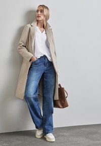 Beige Wollmantel über einer weißen Bluse, kombiniert mit weit geschnittenen blauen Jeans und weißen Turnschuhen. Braune Handtasche an der Seite. Flacher Hintergrund.