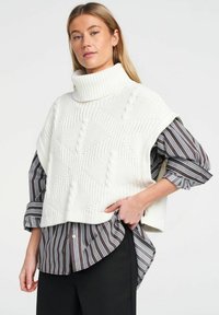 Fehér kötött garbós pulóver mellény, texturált gyémántmintákkal, fekete-fehér csíkos inggel viselve, fekete nadrággal párosítva.