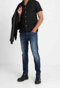 Czarna koszula z krótkim rękawem zapinana na guziki z kieszonką z przodu, niebieskie dżinsy oraz czarne skórzane buty. Model trzyma czarną kurtkę.