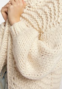 Handen dicht bij de borst, gekleed in een dikke, roomkleurige, grofgebreide trui met gedetailleerde geweven patronen en losse mouwen.