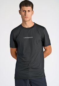 Svart kortärmad skjorta i ett lätt, fuktavvisande tyg med subtil textur och en vit "LINDBERGH"-logotyp på bröstet.