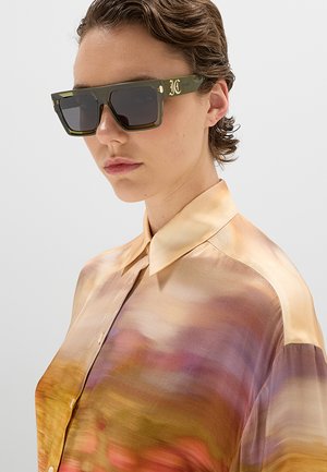 Giovane che indossa occhiali da sole rettangolari verdi e una camicia setosa multicolore con una sfumatura di toni gialli, viola e arancioni.